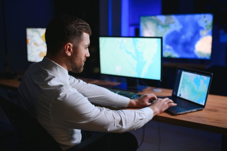 Analyst working late at a desk with monitors showing maps and a laptop